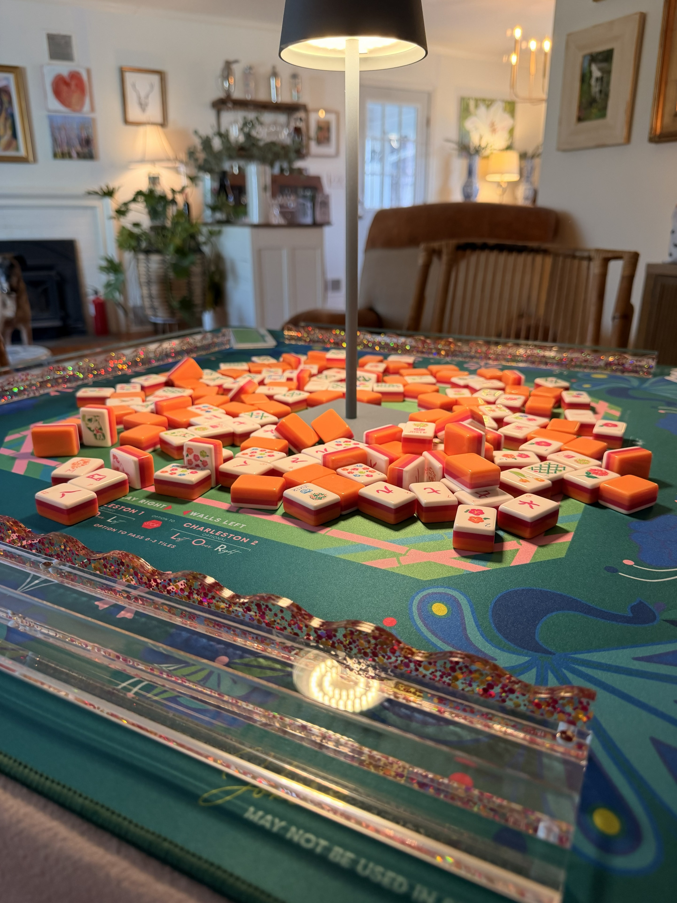 Orange mahjong tiles mid-shuffle on teal floral mat with glitter racks in home interior